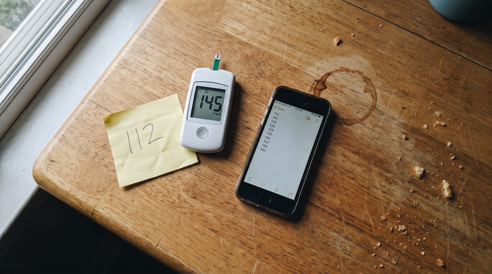 A kitchen table seen from above: a glucose meter reading 145, a sticky note reading 112, and a phone showing a column of numbers